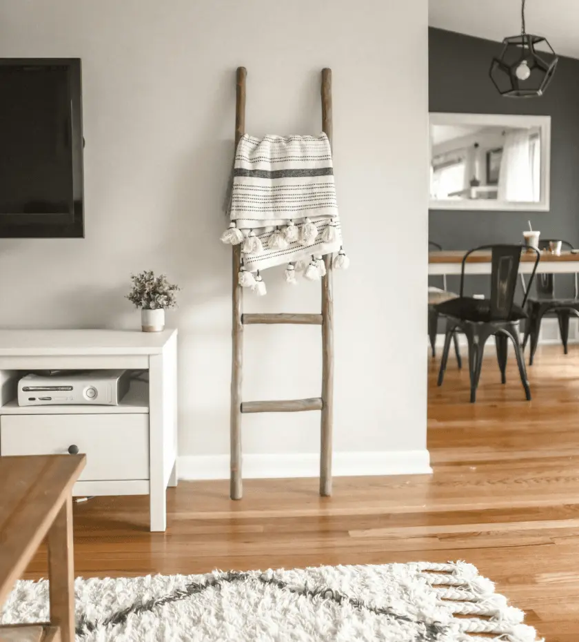 A cozy living space shows a TV mounted above a white media console, a decorative wooden ladder with a tasseled blanket, and a dining area with dark chairs visible in the background.