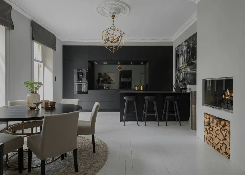 A modern black kitchen and dining area with white floors, a round dining table with chairs, and a geometrically-designed chandelier. There's also a fireplace with stacked logs on the right